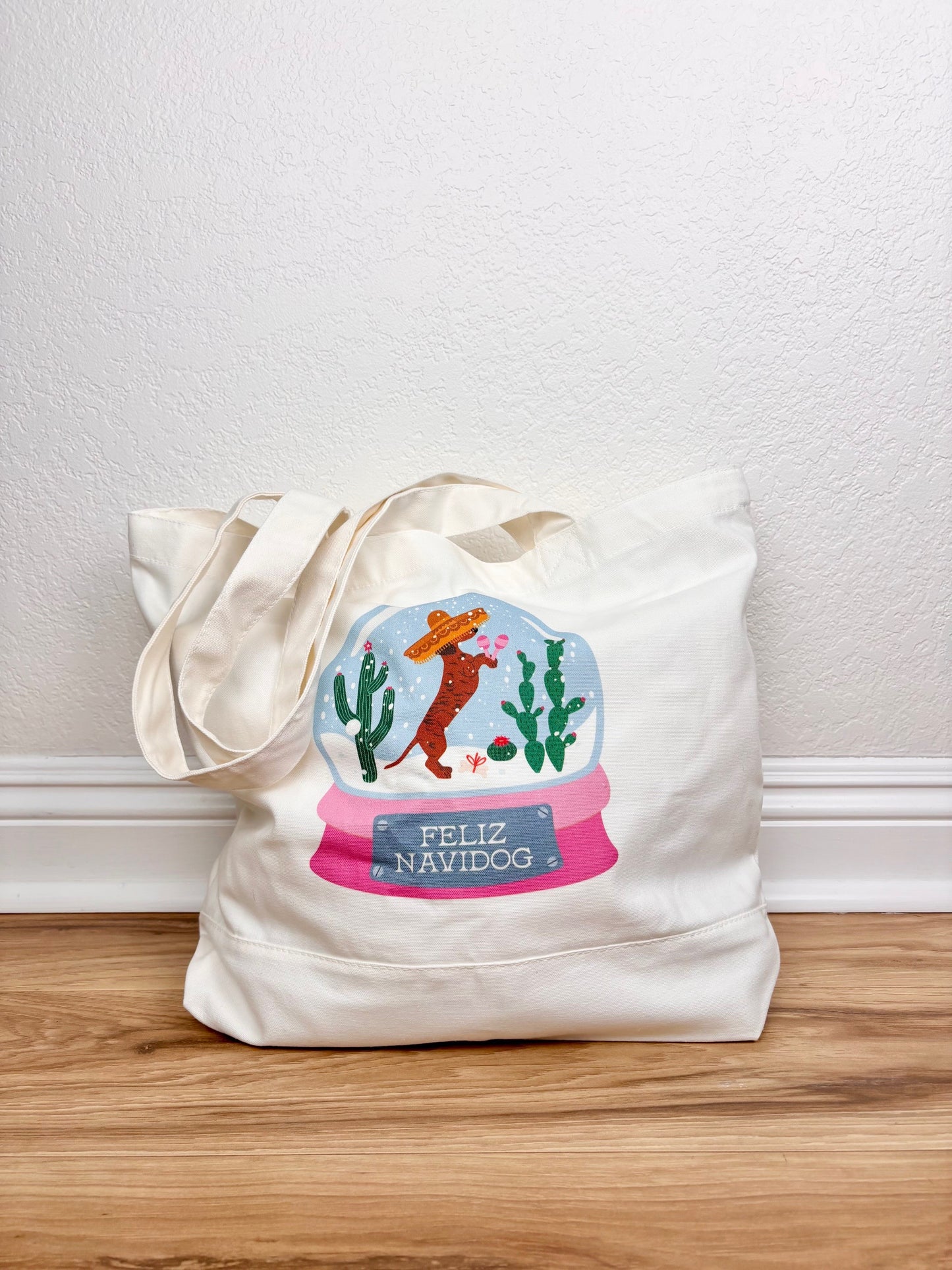 Tote bag with a festive design featuring a snow globe, cactus, and Santa on a wooden surface.