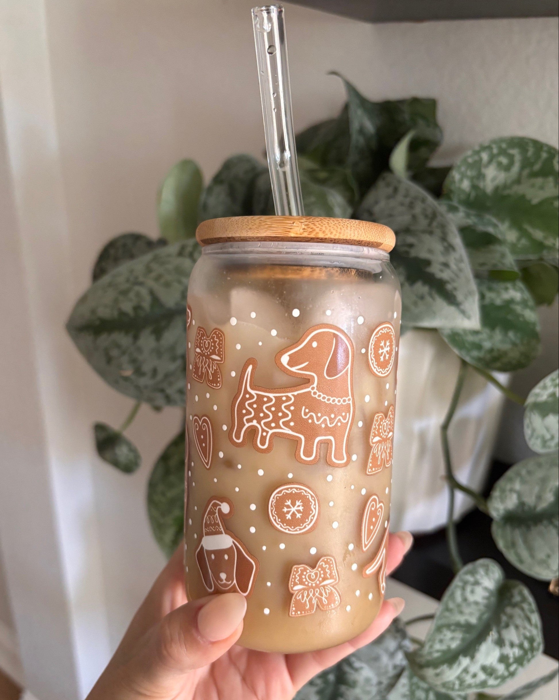 Hand holding a glass tumbler with wooden lid and straw, featuring dog illustrations, in front of green plants.