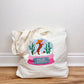 Tote bag with a festive design featuring a snow globe, cactus, and Santa on a wooden surface.