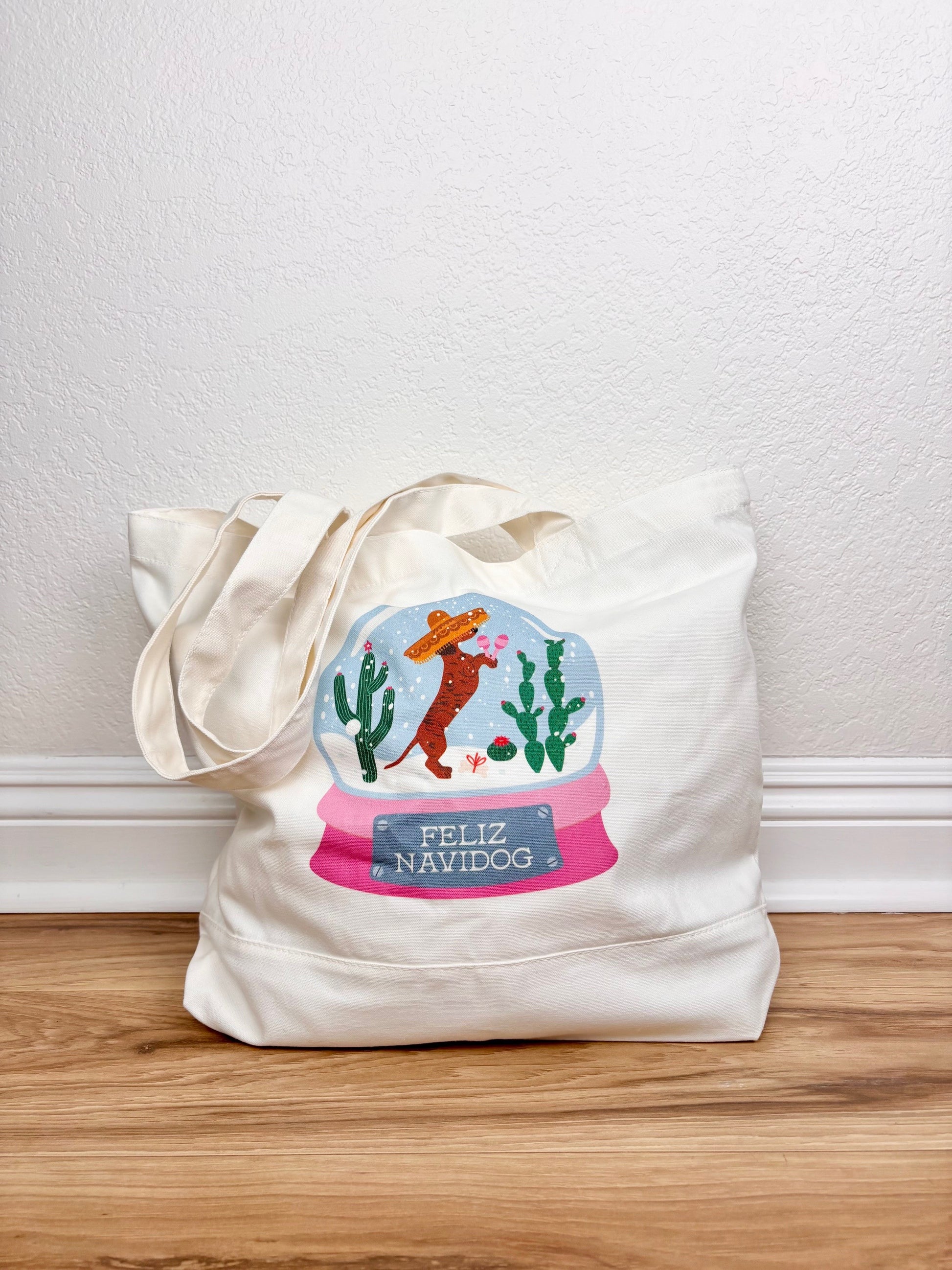 Tote bag with a festive design featuring a snow globe, cactus, and Santa on a wooden surface.
