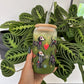A hand holds a clear glass tumbler with a bamboo lid and clear straw, decorated with colorful skeleton dogs and a red heart, filled with an iced green drink. The background features a green patterned houseplant against a white wall.