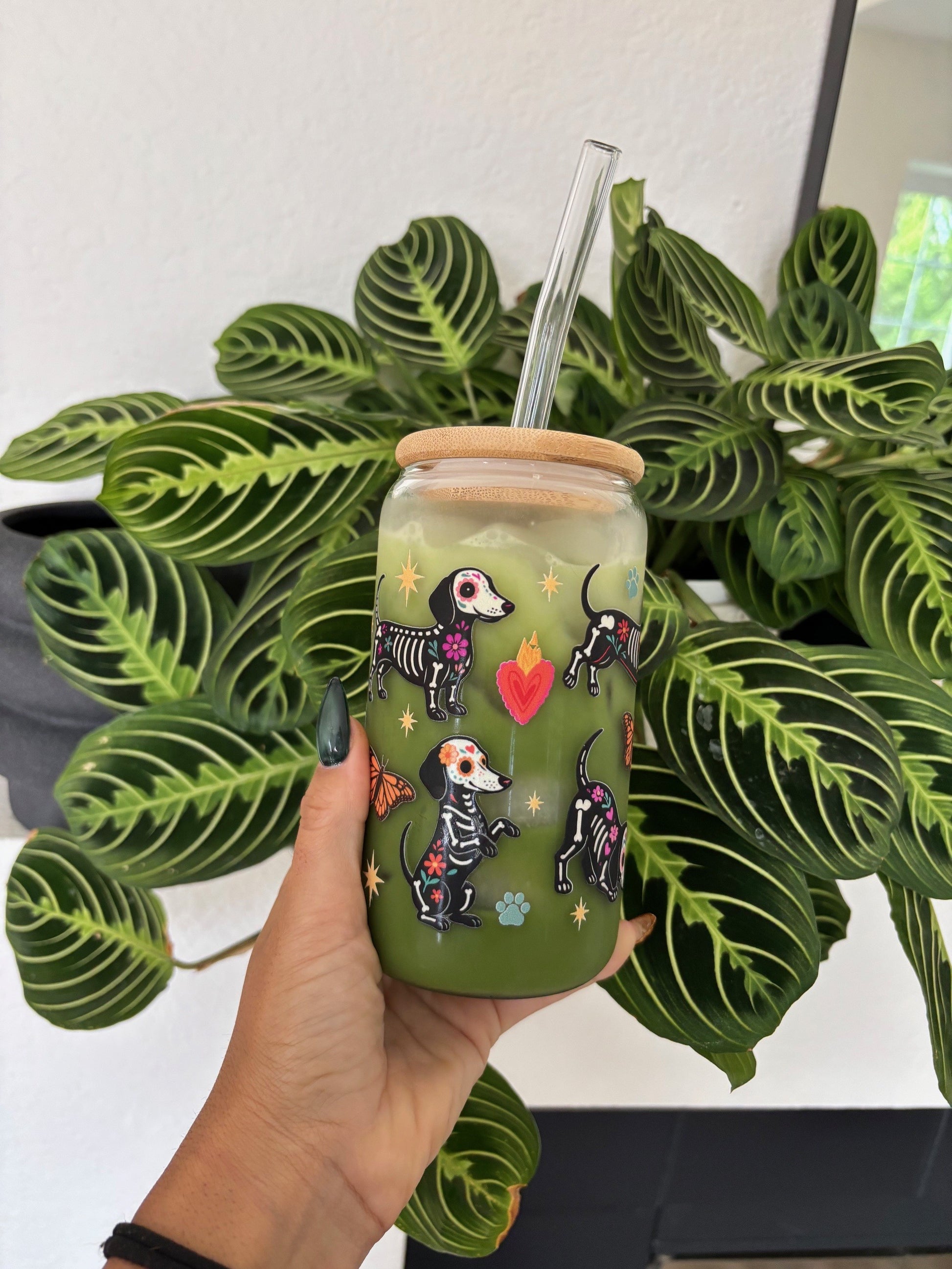 A hand holds a clear glass tumbler with a bamboo lid and clear straw, decorated with colorful skeleton dogs and a red heart, filled with an iced green drink. The background features a green patterned houseplant against a white wall.