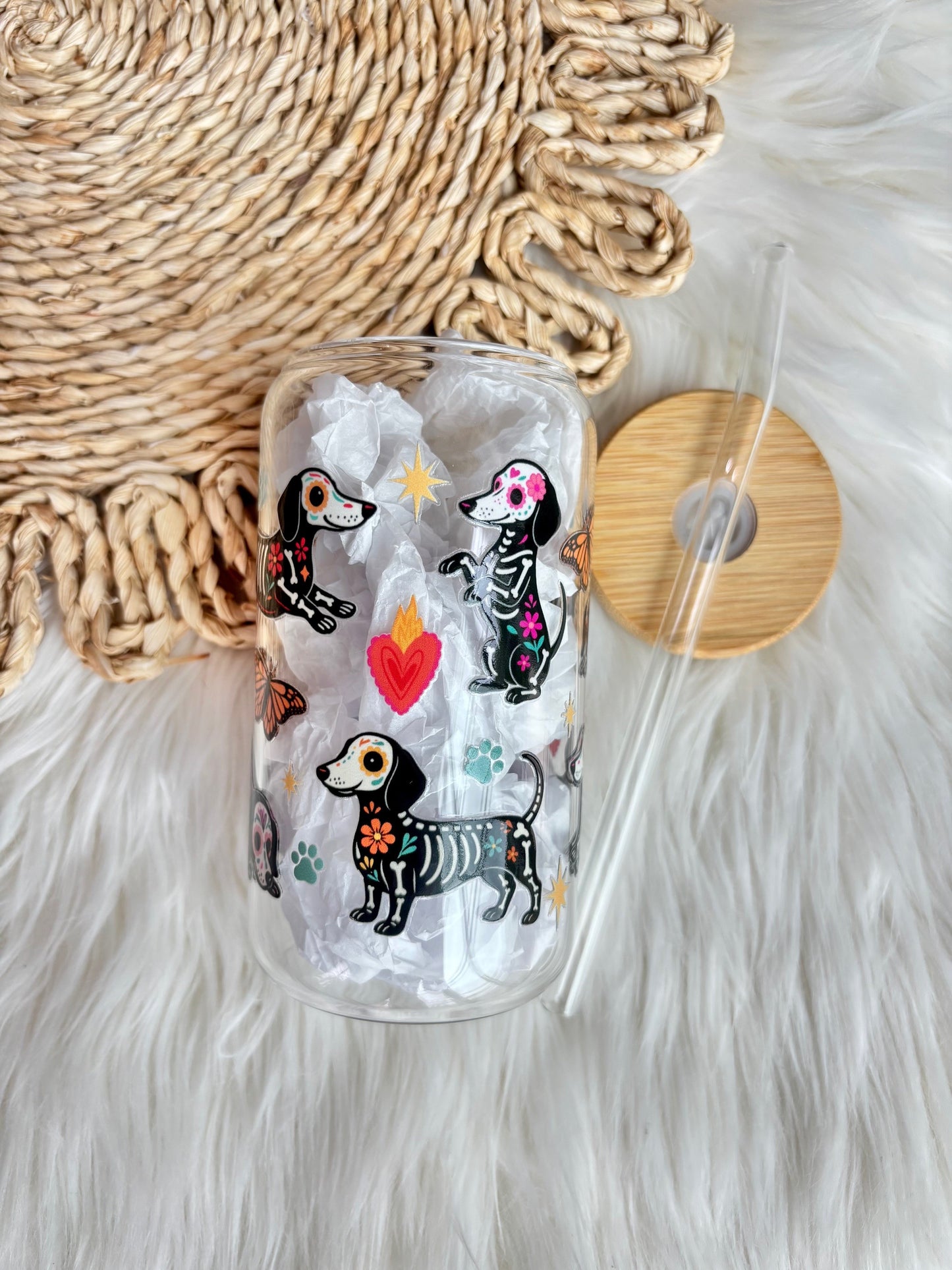 A clear glass can-shaped tumbler with a bamboo lid and straw, decorated with colorful dachshund illustrations in a Day of the Dead style, resting on a white faux fur surface next to a woven mat.