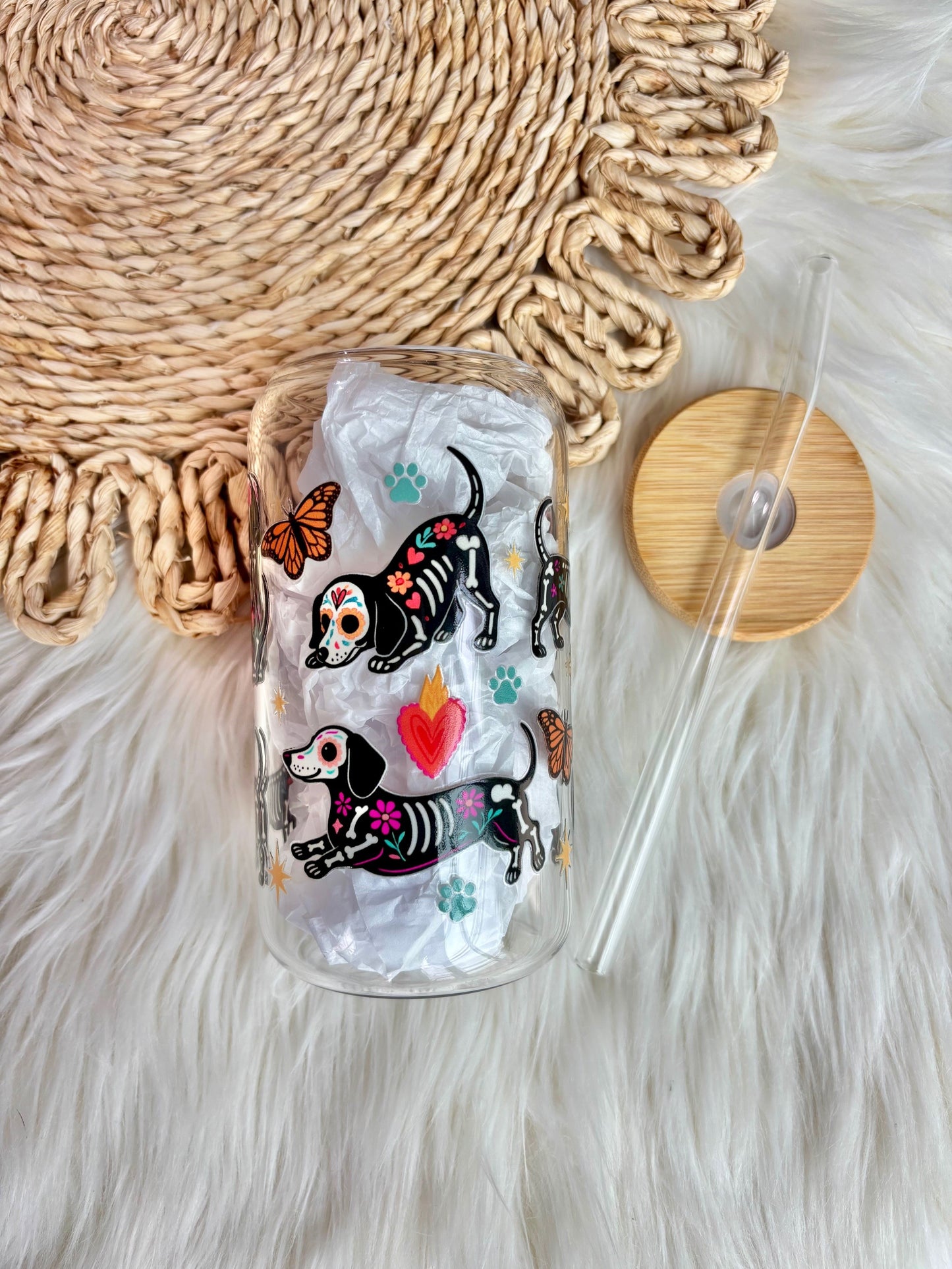 A clear glass tumbler with a bamboo lid and clear straw, decorated with colorful dachshund illustrations and paw prints, displayed on a white faux fur surface next to a woven mat.