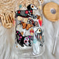 A clear glass tumbler with a straw, decorated with colorful illustrations of dachshund dogs in Day of the Dead style, an orange butterfly, and small floral motifs, shown on a white faux-fur surface with a woven mat and wooden lid in the background.