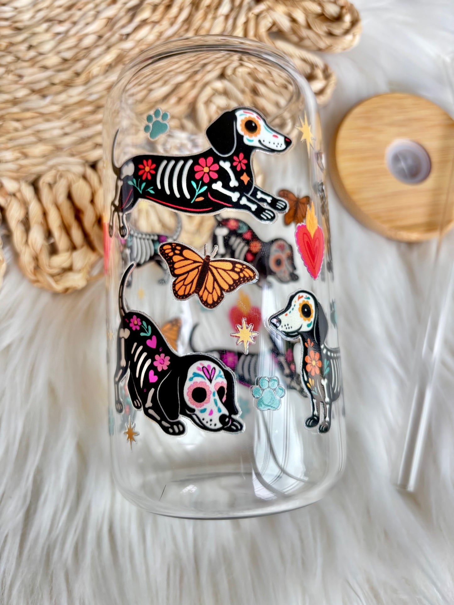 A clear glass tumbler with a straw, decorated with colorful illustrations of dachshund dogs in Day of the Dead style, an orange butterfly, and small floral motifs, shown on a white faux-fur surface with a woven mat and wooden lid in the background.