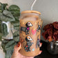A hand holds a clear glass iced coffee tumbler with a bamboo lid and glass straw. The clear glass is decorated with black dachshund skeleton illustrations, pink hearts, and small colorful butterfly and paw print accents.