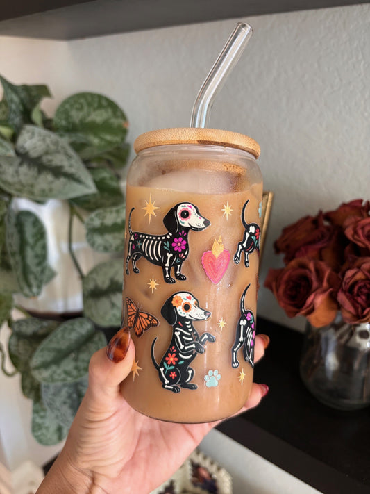 A hand holds a clear glass iced coffee tumbler with a bamboo lid and glass straw. The clear glass is decorated with black dachshund skeleton illustrations, pink hearts, and small colorful butterfly and paw print accents.